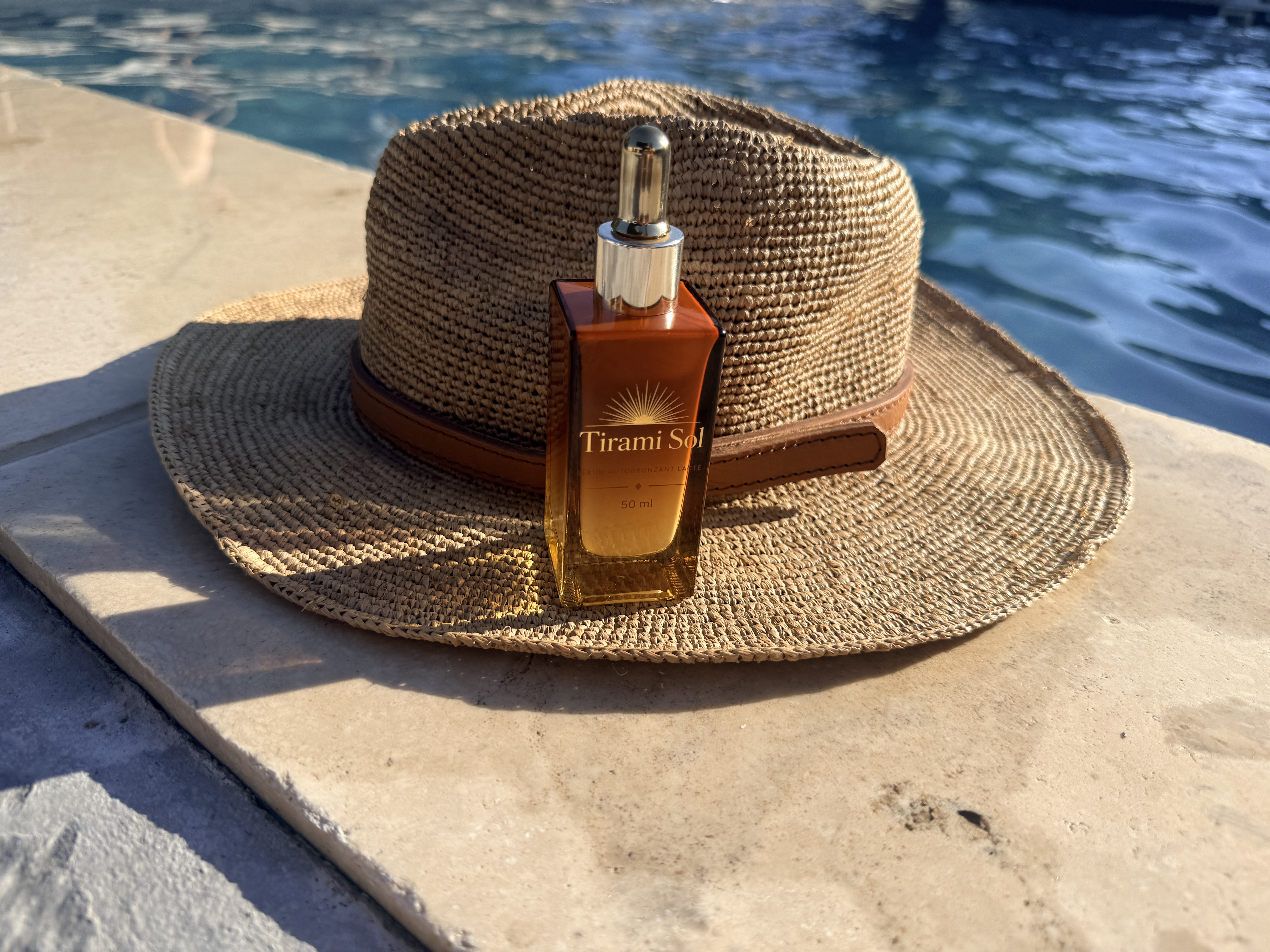 Photo du véritable flacon de sérum autobronzant TiraMiSol 50 ml devant un chapeau, au bord de la piscine